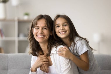 Neşeli genç anne tatlı kızı sırtında taşıyor, gülümsüyor, gülüyor, anneliğin tadını çıkarıyor. Mutlu anne ve çocuk, evde kanepede eğleniyor, aile fotoğrafı için kameraya bakıyor.