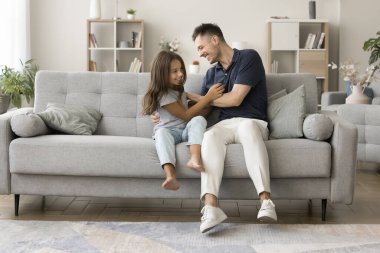 Mutlu, genç baba yumuşak, şık koltukta tatlı kızı gıdıklıyor, rahat oturma odasında çocukla aktif oyun oynuyor, kıza sarılıyor, gülüyor, eğleniyor, eğleniyor, eğleniyor.
