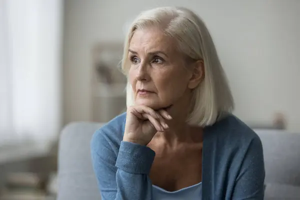 stock image Pensive old woman sits at home alone, look aside, deep in thoughts about health problems, feel anxious, suffer from sadness, try to find answers to issues. Melancholy, loneliness, life on retirement