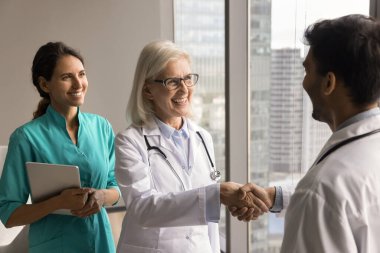 Genç erkek doktorla tokalaşan, gülümseyen, gülen, yeni sağlık uzmanı işe alan, genç meslektaşlarına şükranlarını sunan, kıdemli klinik patronu mutlu kadın.