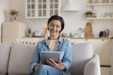 Dost canlısı, serbest çalışan bir kadın uzaktan kumanda için tablet kullanıyor, evden çalışıyor. Olgun bir bayanın cihaz yazması, evde boş vakitlerin tadını çıkarması, internetten kitap okuması, görüntülü konuşma.