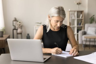 Ciddi, olgun, emekli bir kadın, dizüstü bilgisayarda ders çalışıyor, not defterine notlar yazıyor, internetteki eğitim ağlarını, profesyonel kursları izliyor, sesli dersler dinliyor.