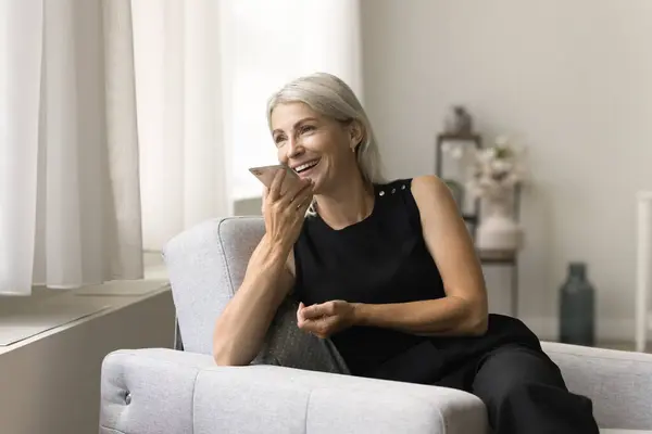 stock image Cheerful blonde senior woman enjoying leisure, mobile phone communication in cozy armchair, talking on smartphone on speaker, holding cell at face, smiling, laughing, having fun, looking away