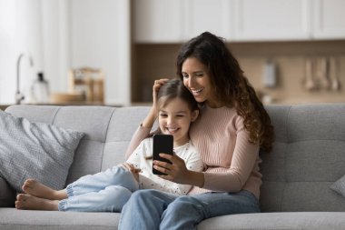 Neşeli, güzel, siyah saçlı anne ve pozitif kız çocuğu akıllı telefondan video konuşmaları, ev kanepesinde dinlenmeleri, cep telefonlarını internet üzerinden aile iletişimi için kullanmaları.