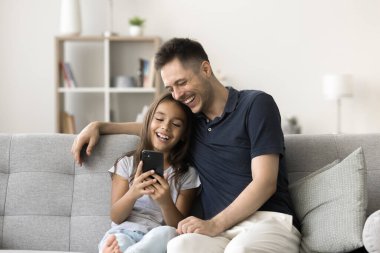 Mutlu baba ve küçük kızı akıllı telefondan online iletişimi kullanıyorlar, dijital aygıtı birlikte kullanıyorlar, evde kanepede dinleniyorlar, boş zamanların, ilişkilerin, babalığın tadını çıkarıyorlar.
