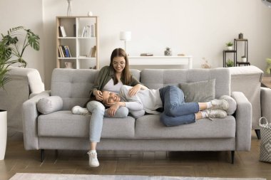 Mutlu anne ve genç kız modern ev içinde rahat bir kanepede dinleniyor. Pozitif gevşemiş genç anne, kafası çocukların kucağında kanepede uzanıyor, kızla sohbet ediyor, gülüyor.