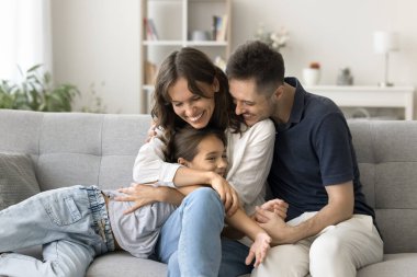 Mutlu, genç ebeveynler rahat bir ev kanepesinde tatlı şirin küçük kıza sarılıyorlar, çocuğa sevgiyle sarılıyorlar, hayranlıkla sarılıyorlar, gülüyorlar, ailecek kızlarıyla vakit geçiriyorlar.