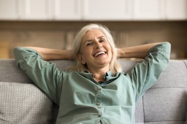 Hoş, güzel, sarışın, yaşlı bir kadının ev portresi rahat koltukta dinleniyor, dirsekleri açık yumuşak sırtına yaslanıyor, vücudunu esnetiyor, temiz hava soluyor, dişlek tebessümle kameraya bakıyor.