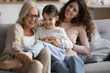 Mutlu bir büyükanne ve anne kanepenin üzerinde neşeli bir kızla oynuyor, gülen çocuğu gıdıklıyor, eğleniyor, aile aktivitelerinin keyfini çıkarıyor, yakınlık, bağ kurma, oyun zamanı.