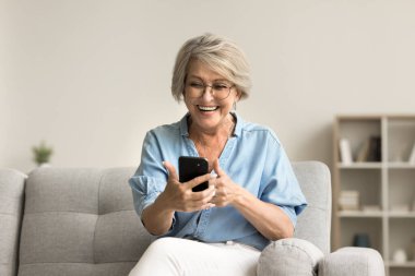 Neşeli, olgun, emekli bir kadın, mutlu haberlerle mesajlar okuyor, cep telefonu tutuyor, gülümsüyor, gülüyor, video konuşmaları yapıyor, evdeki kanepede kablosuz bağlantının keyfini çıkarıyor.