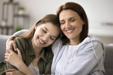 Mutlu barışçıl anne ve güzel genç kız evdeki kanepeye oturuyor, kapalı gözlerle sarılıyor, gülümsüyor, gülüyor, konuşuyor, sağlıklı aile ilişkilerinden keyif alıyor.