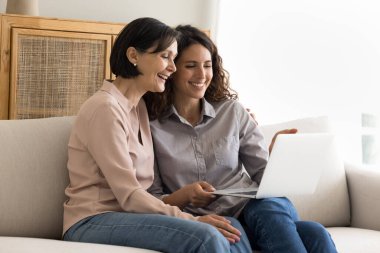 Mutlu çoklu nesiller ailesi internette eğleniyor, genç yetişkin kızı yetişkin annesi komedi filmi seyrediyor, kanepede dizüstü bilgisayarla oturuyor, videoların keyfini çıkarıyor, boş zamanlarında evde dinleniyor.