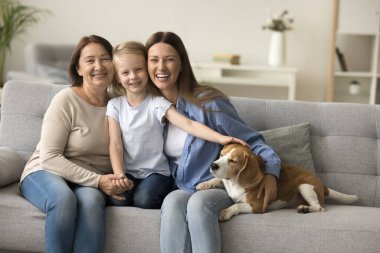 Anaokulunda tatlı bir kız çocuğu kanepesinde sevimli bir av köpeğini okşuyor, sevgi dolu anne ve büyükannesiyle oturuyor, kameraya bakıyor, gülümsüyor. Neşeli güzel kızlar ve üç nesil aile portresi