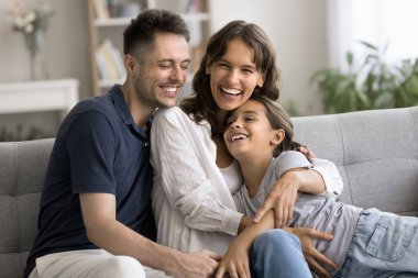Heyecanlı genç anne ve baba, küçük kızına evde kanepede sarılıyor, sevgiyle, ilgiyle, gülümsemeyle, gülümsemeyle, kameraya bakarak, ebeveynlikten keyif alarak, boş zamanlarında,