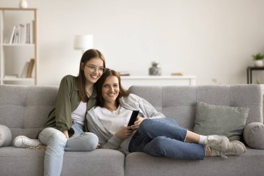 Neşeli anne ve güzel genç çocuk rahat bir evde rahatlıyor, internet iletişimi için cep telefonunu kullanıyor, dijital aletlerle kanepede dinleniyor, boş vakitlerin tadını çıkarıyor, kameraya bakıyor.