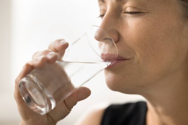 Mutlu, dingin, olgun bir bayan, kapalı gözlerle maden suyu içiyor, şeffaf camlar tutuyor, kuru cilt bakımı için su dengesini sağlıyor, sağlıklı metabolizma, gülümsüyor.