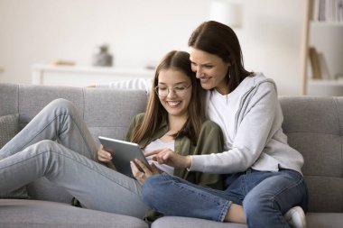 Mutlu anne ve ergen kız birlikte alışveriş yapmak için online başvuru kullanıyorlar, kanepede dinleniyorlar, dijital tablet bilgisayar tutuyorlar, gülümsüyorlar, gülüyorlar, evcil internet iletişiminden keyif alıyorlar.