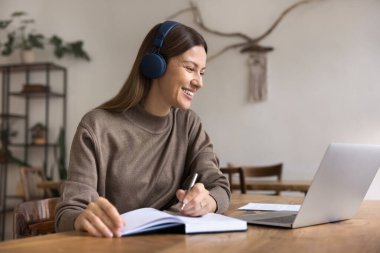 İnternette okuyan mutlu yetişkin kadın öğrenci, dizüstü bilgisayar, kablosuz kulaklık kullanıyor, video konferans görüşmesinde konuşuyor, webinar izliyor, notlar yazıyor, gülüyor, gülüyor.