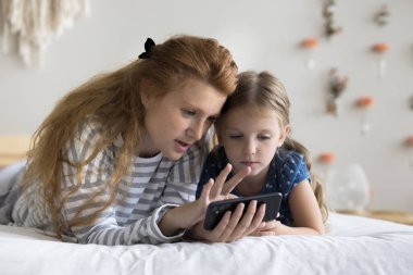 Odaklanmış genç bir anne, sevimli bir çocuğa cep telefonuyla eğitim uygulamasını kullanmayı öğretiyor, dijital aletlerle yatakta dinleniyor, çevrimiçi oyun oynuyor, internet hizmeti veriyor.