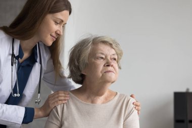 Olumlu güzel doktor yaşlı, düşünceli, kayıp hasta bayanı görüyor, düşünceli, yaşlı bir kadının omuzlarına dokunuyor, başka tarafa bakıyor, gülümsüyor, tıbbi yardım yapıyor, destek oluyor.
