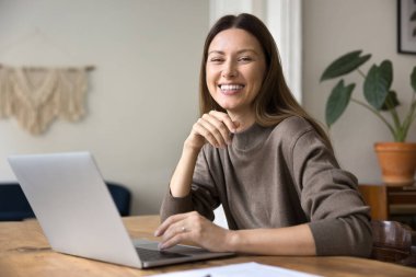 Ev bilgisayarında çalışan başarılı girişimci kadın, mükemmel dişli gülümsemeyle fotoğraf için kameraya bakıyor, çalışma masasında poz veriyor, uzaktan işten zevk alıyor, internet üzerinden iş yapıyor.