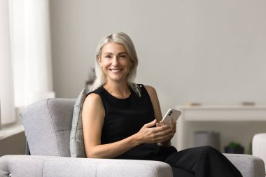 Neşeli, çekici, olgun bir kadın, elinde beyaz bir akıllı telefon tutuyor, rahat koltukta dinleniyor, kameraya bakıyor, portre için gülümsüyor, kullanışlı internet uygulamalarını destekliyor,