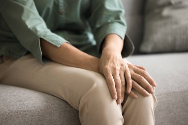 Kanepede rahat rahat oturan, kollarını kavuşturan, bileklerini dizlerinin üstüne koyan, dirseklerini kalçalarına dayayan, duruşunu kapalı tutan, vücut hareketi yapan yaşlı bir kadın, endişe. Kadın elleri yakın.