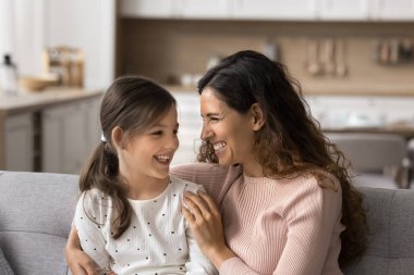 Heyecanlı güzel anne ve pozitif küçük kız evdeki kanepede eğleniyor, konuşuyor, gülüyor, yakınlık ve güven hissediyor. Mutlu anne koltukta çocuğuna sarılıyor, aile tatilinin tadını çıkarıyor.