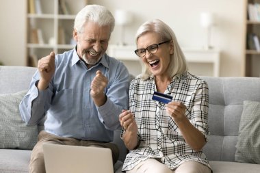 Mutlu yaşlı çift internet alışverişi için dizüstü bilgisayar ve kredi kartı kullanıyor. Kazanan ödül, indirim, hediye, kazancı, geliri, başarıyı kutluyor. Evet el hareketi yapıyor, gülüyor.