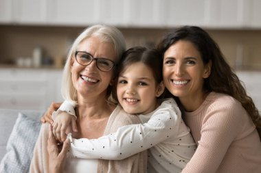 Neşeli, çekici büyükanne, anne ve torun, sevgiyle kucaklaşan, yakın ilişkiden zevk alan, aile eğlencesi, mükemmel dişlek gülümsemeyle kameraya bakan.