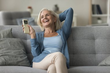 Mutlu, orta yaşlı bir kadın uzaktan kumandayı tutuyor, kanepede oturan klimayı açıyor, oturma odasındaki temiz havanın tadını çıkarıyor. Akıllı ev sahibi, rahat yaşam, iklim kontrolü