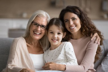 Mutlu anne ve yaşlı büyükanne, tatlı ergen kızı kucaklıyor, aile portresi için koltukta poz veriyor, kameraya dişlek tebessümle bakıyor, aile bağlarının keyfini çıkarıyor, birlikte vakit geçiriyorlar.