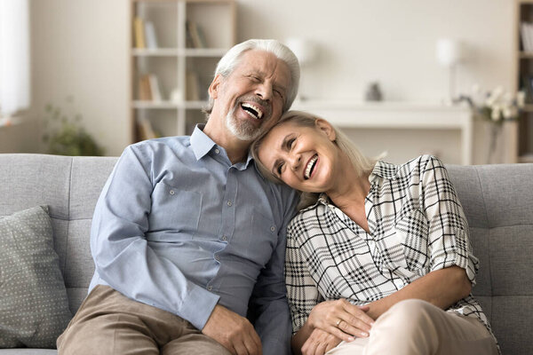 Счастливая довольно зрелая жена отдыхает на диване с головой на плече мужа. Старшие семейные пары отдыхают на диване вместе, разговаривают, обсуждают отпуск, улыбаются, смеются