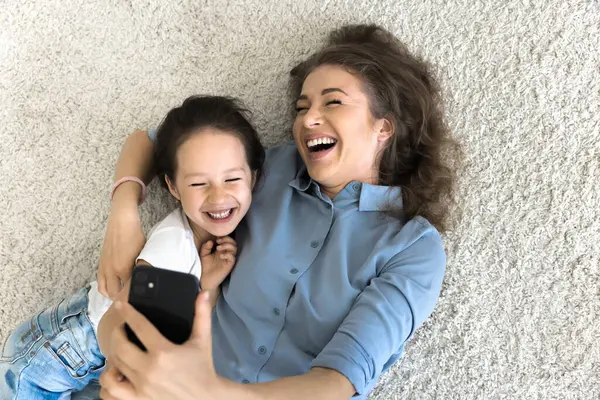 Neşeli genç bir anne, mutlu tatlı küçük bir kıza sarılıyor, yumuşak halıda dinleniyor, sıcak zeminde, cep telefonunda selfie çekiyor, sırtüstü uzanıyor, gülüyor, gülüyor, aile içi internet iletişiminden keyif alıyor.