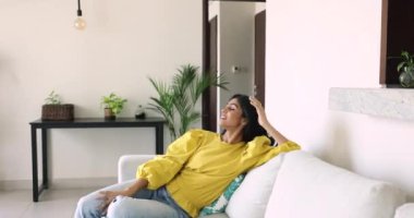 Çekici mutlu Hintli kadın gündelik kıyafetlerle modern aydınlık oturma odasına giriyor rahat bir kanepeye oturuyor ve hafta sonları evde yalnız başına keyfine bakıyor. Tatil, konaklama, izin günü ve hayal.