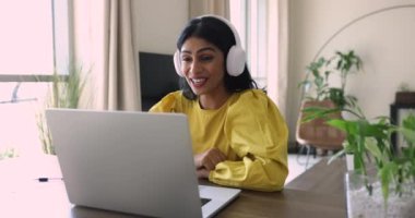 Hintli genç bir kadın masasında oturuyor, modern kulaklıklar takıyor, laptop ekranına bakıyor, görüntülü konuşuyor, uzaktan çalışıyor, özel online dersler veriyor, danışmanlık yapıyor, destek veriyor. Webinar, çevrimiçi iletişim