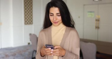 Çekici Latin kadın cep telefonu tutuyor, mesaj okuyor, iyi haberler alıyor, gülümsüyor, sosyal ağda vakit geçirmekten hoşlanıyor. Pozitif kadın akıllı telefondaki yeni uygulamayı kullanarak evde e-randevu hizmetlerinden hoşlanıyor