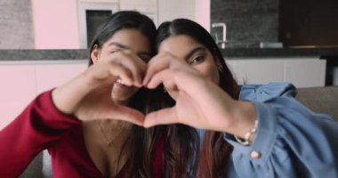 İki Hintli genç kadın gülümsüyor kameraya bakıyorlar, parmaklarını birleştiriyorlar, kalp sembolü gösteriyorlar, aşka işaret ediyorlar, mutlu yüzleri kapatıyorlar. Anlayış, dostluk, aile bağları, birlik