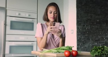 Modern akıllı telefon tutan çekici genç bir kadın sağlıklı sebze salatası hazırlığından dikkati dağılmış. Yemek blogları, çevrimiçi yemek kitabı tarifleri başvuru kullanım, mutfakta yemek pişirirken sohbet