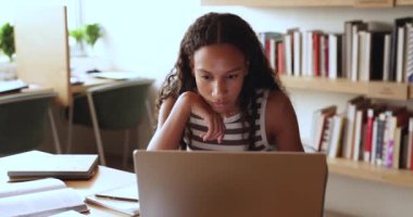 Odaklanmış Afrikalı genç kız kütüphanede masada oturuyor, laptop ekranına bakıyor, webinar izliyor, proje üzerinde çalışıyor, mesaj okuyor, internette arama yapıyor. E-öğrenme, eğitim, modern teknoloji