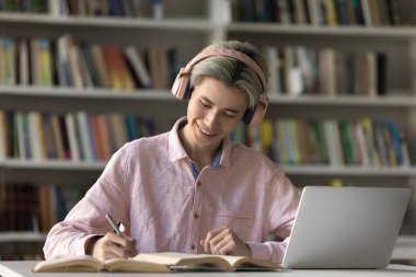 Pembe kablosuz kulaklıklı neşeli Asyalı öğrenci kütüphanede ders çalışmaktan, internetten ders dinlemekten, dizüstü bilgisayarda web sitesinden not yazmaktan, makale yazmaktan, rapor yazmaktan, kitaplarda çalışmaktan keyif alıyor.