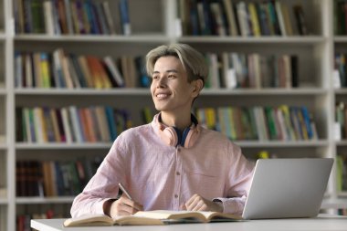 Kolej kütüphanesinde ödev yapan neşeli Asyalı öğrenci, dizüstü bilgisayara notlar yazıyor ve kitapları açıyor, başka tarafa bakıyor, makale için eleştirel fikirler düşünüyor, gülümsüyor, gülüyor.