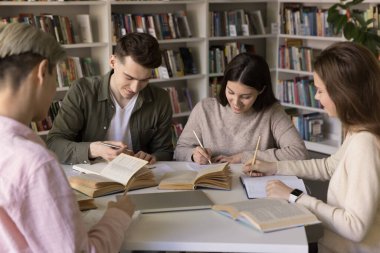 Bir grup pozitif, çalışkan, zeki öğrenci kütüphanede çalışıyor, açık kitapların başında birlikte oturuyor, notlar yazıyor, makale yazıyor, konuşuyor, takım çalışması yapıyor, araştırma yapıyor, rapor hazırlıyor.