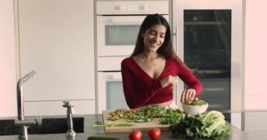 Mutlu genç Hintli kadın yemek pişiriyor, evde dans ediyor. Güzel bir kadın diyet salatası için sebze kesiyor, favori parçayı dinliyor, müziğe geçiyor, tasasız görünüyor mutfakta yemek hazırlıyor.