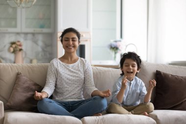 5 'lik oğlu olan gülümseyen Hintli anne koltukta birlikte meditasyon yapıyor, rahat koltukta oturuyor, küçük çocuğuna yoga yapmayı öğreten mutlu bir anne, sağlıklı bir yaşam tarzı, boş vakitlerin tadını çıkarıyor.