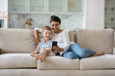 Mutlu Hintli anne ve Kafkas 7 'nin oğlu akıllı telefonu birlikte kullanıyor, rahat rahat koltukta oturuyor, çocuklu gülümseyen bir anne telefon ekranına bakıyor, çizgi film izliyor, eğleniyor.