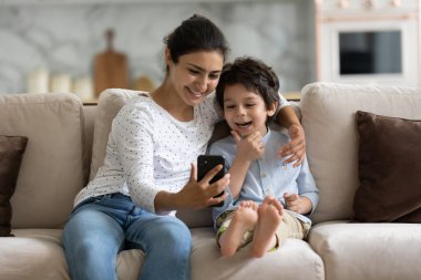 Gülümseyen Hintli anne ve oğul akıllı telefonla eğleniyor, sarılıyor, rahat koltukta oturuyor, 5 'li yaşlarda bir çocukla mutlu bir anne telefon ekranına bakıyor, çizgi film izliyor ya da video görüşmesi yapıyor.