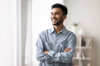 Young Indian man spend time at home, standing with arms crossed on chest smile, look away, enjoy day-off at own or rented apartment, rest, relish pleasant thoughts, good life opportunity and leisure clipart