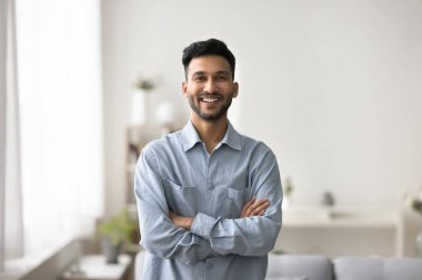 Portrait of happy Indian man having wide toothy smile, pose with arms-crossed, look at camera, spend carefree time in modern living room, enjoy day-off. Profile picture of homeowner, renter or tenant clipart