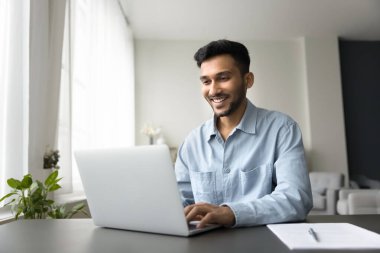 Gülümseyen genç serbest çalışan adam evde uzaktan çalışmaktan, dizüstü bilgisayarında e-posta yazmaktan, iş arkadaşlarıyla internetten konuşmaktan, internete bakmaktan hoşlanıyor. Tatmin olmuş Hintli erkek işçi, rahat tele-iş için modern cihaz kullanıyor.